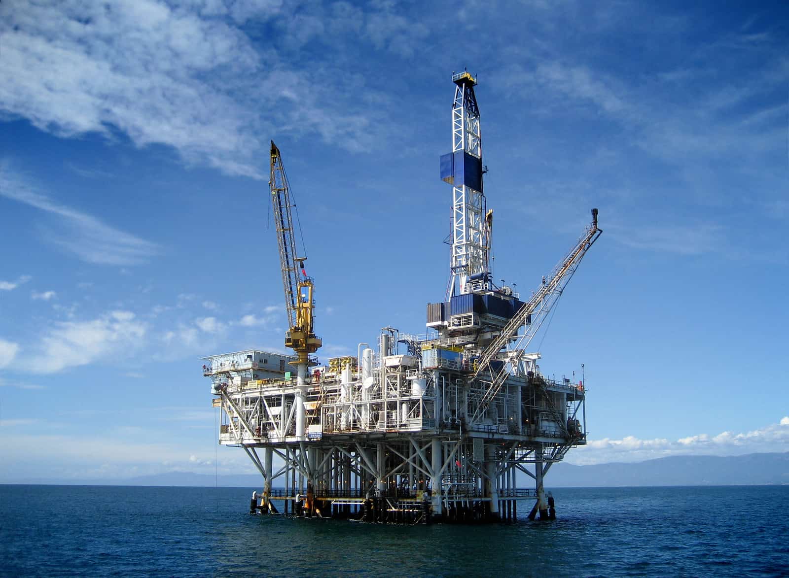 An offshore oil rig platform in the middle of the ocean against a backdrop of blue sky and clouds. The Oil & Gas platform has various machinery and equipment, as well as cranes extending from it. The calm sea surrounds the structure, dispelling myths about Oil & Gas with its serene presence.
