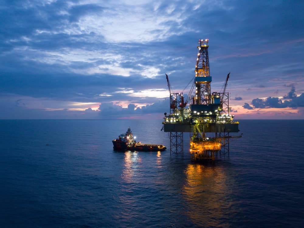 An offshore oil rig is illuminated as it stands tall against a backdrop of a dusky sky and calm sea. A supply vessel, essential for valve maintenance, is positioned near the rig, both reflecting lights on the water's surface. The horizon shows a mix of dark clouds and the fading light of sunset.