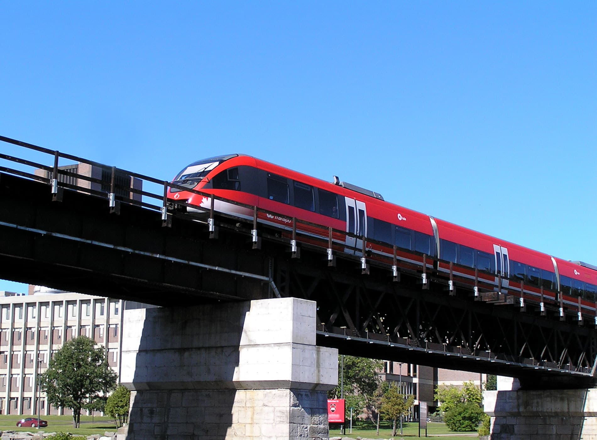 A red passenger train crosses a black metal bridge on a sunny day. Supported by sturdy stone pillars, the rail track glistens from recent Rail Track Maintenance. In the background, buildings and trees with green foliage stand under a clear blue sky.