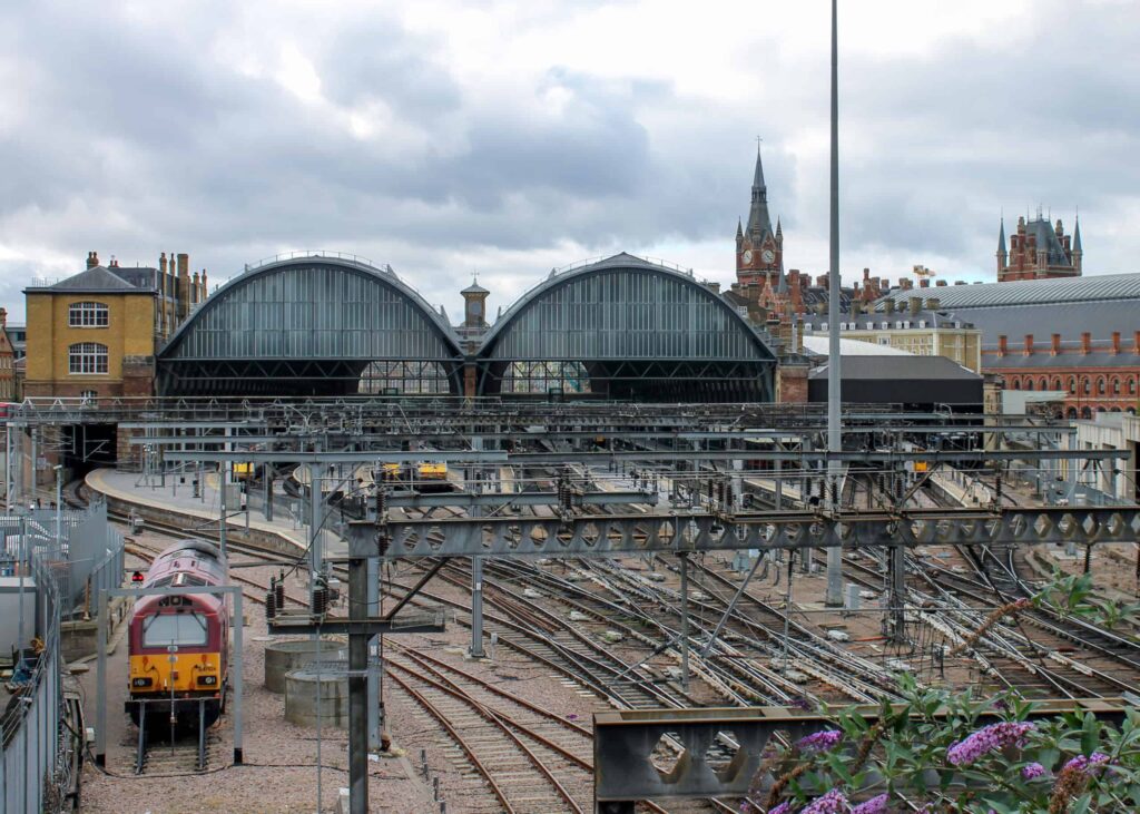 A busy train station with multiple tracks leading to a large building with two arched roofs. Several trains, including a red locomotive, are visible. In the background, a clock tower and various brick buildings stand under a cloudy sky, testaments to curved track maintenance ensuring smooth operation.