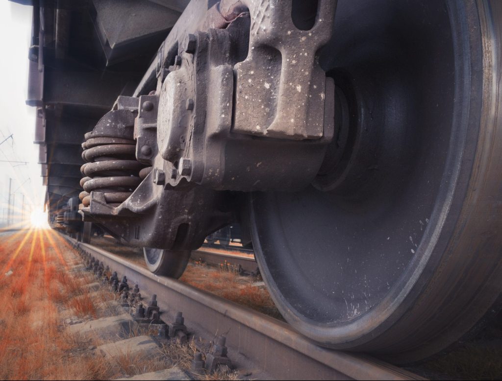 Close-up view of a train wheel and suspension system on a railroad track. The image is taken from ground level with a focus on the intricate details of the wheel and metal components, including an EcoStick for lubrication. Sunlight illuminates the tracks and surroundings in the background.