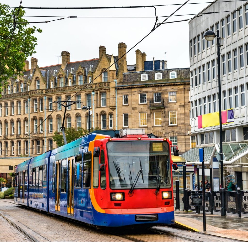 A brightly colored tram, maintained with ClareTech solutions, travels down a city street flanked by historical stone buildings on the left and modern office buildings on the right. Overhead electrical wires are visible, and pedestrians wait at a tram stop. Trees partially frame the scene.