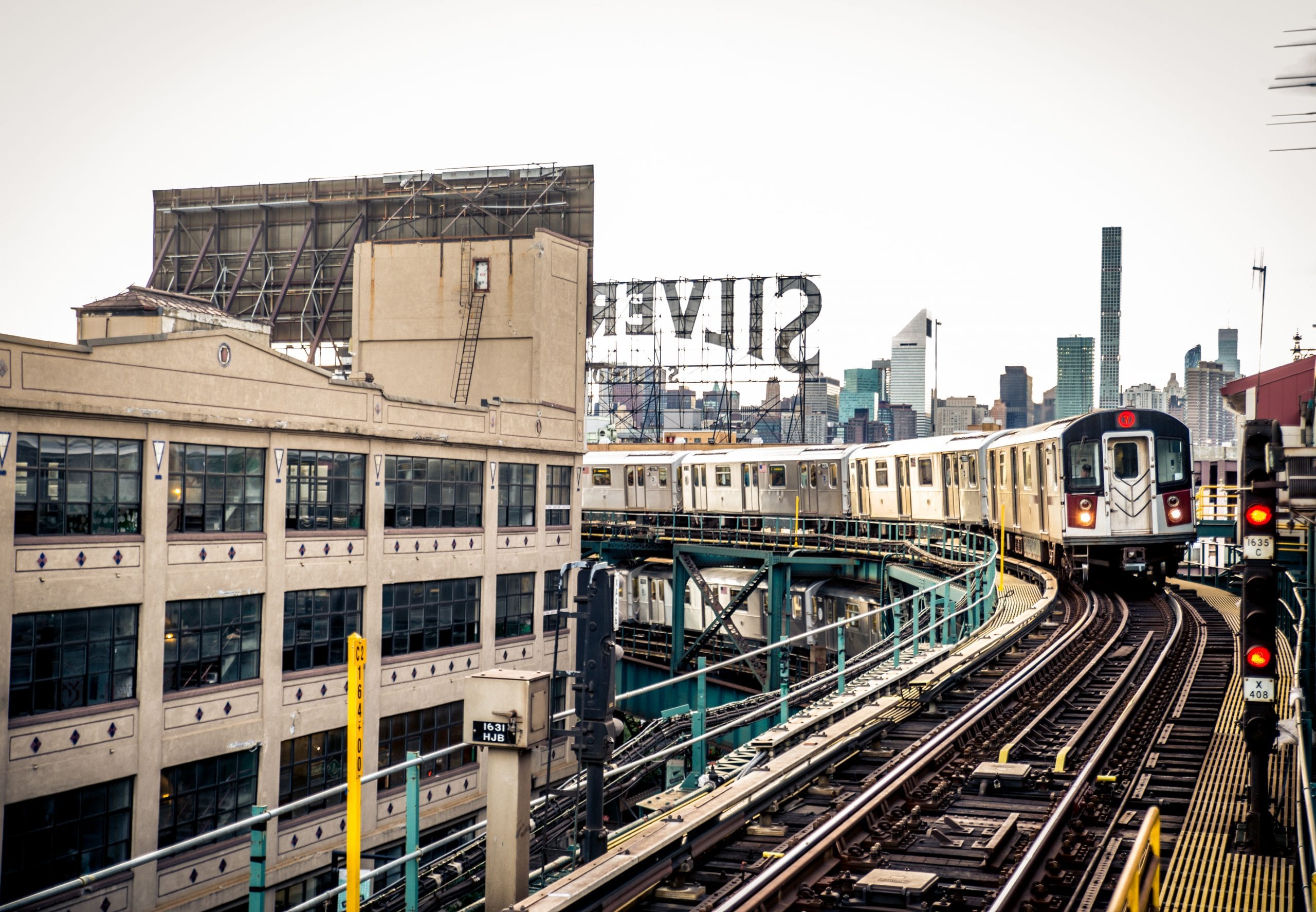 An elevated train with silver and white cars travels along curved tracks past an industrial-looking building in a cityscape. The background includes various skyscrapers, the recognizable Silvercup Studios sign, and features Ecocurve Trackside Grease for a more environmentally friendly commute.